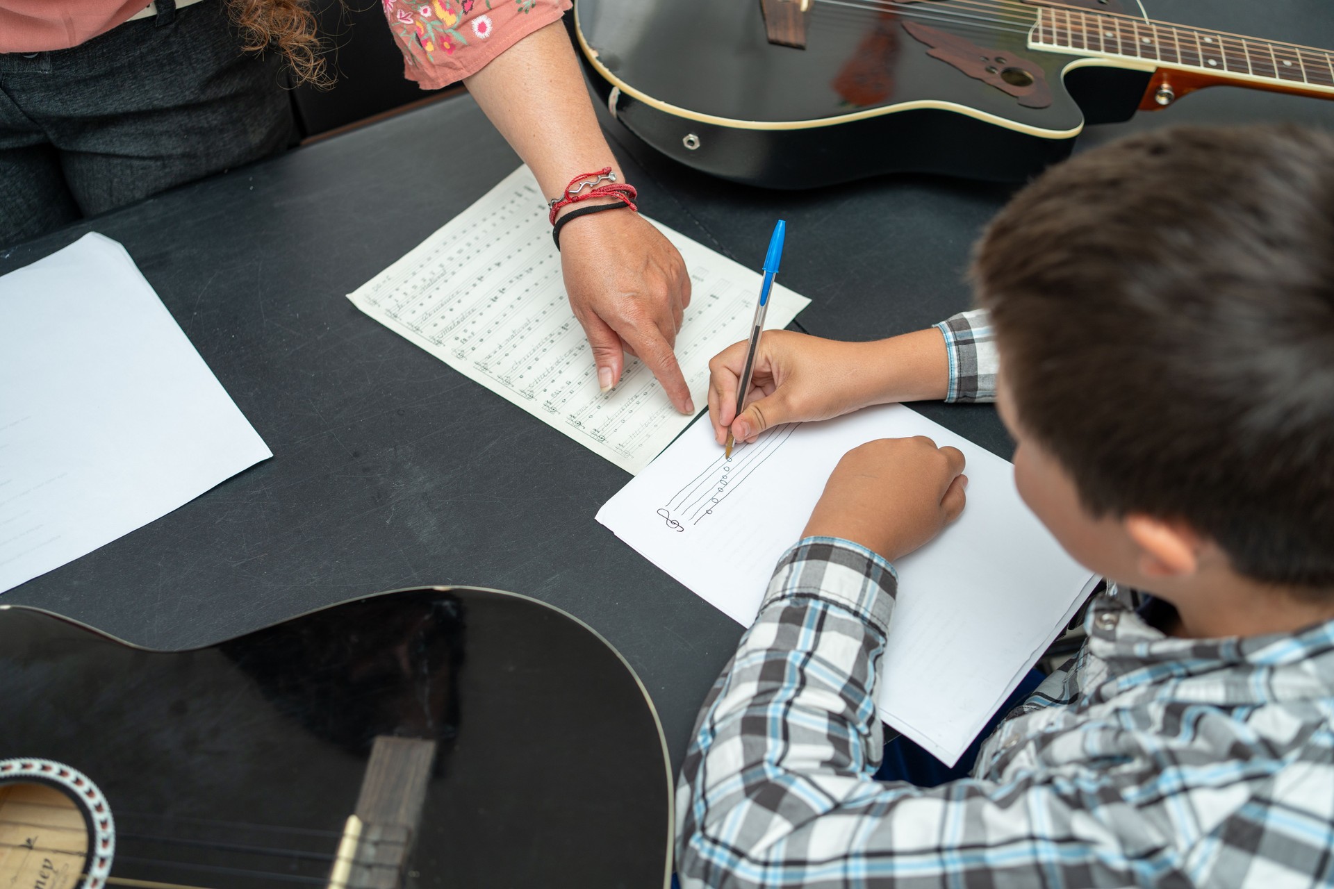 Child writing music notes beside guitar teacher during a private